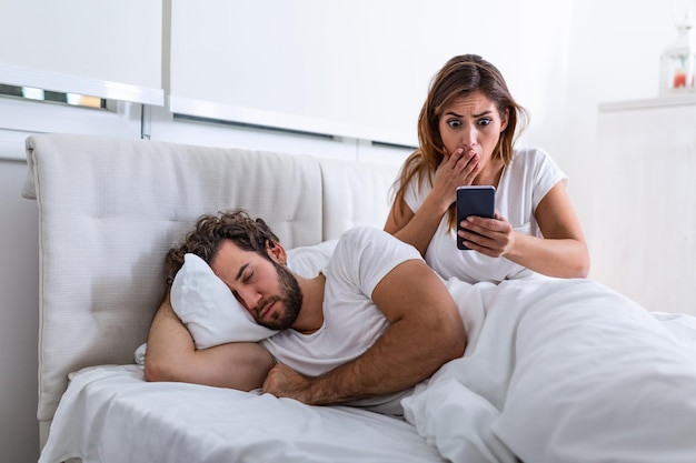 Zazdrosna żona podgląda telefon swojego partnera, gdy ten śpi w łóżku w domu. Zszokowana zazdrosna żona podgląda telefon męża, gdy mężczyzna śpi w łóżku w domu