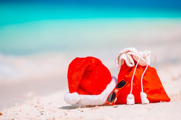 Zdjęcie zbliżenie czerwony worek świętego mikołaja i kapelusz świętego mikołaja na plaży. boże narodzenie podróży wakacje i koncepcja podróży cuprise. akcesoria plażowe z santa hat na białej tropikalnej plaży