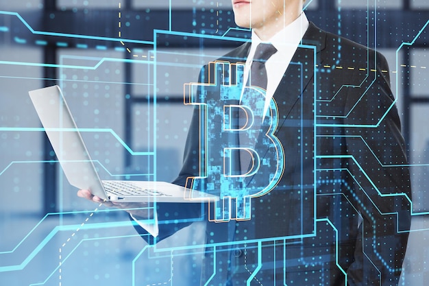 Zbliżenie dłoni biznesmena za pomocą laptopa ze świecącym liniowym hologramem bitcoin na rozmytym tle wnętrza biura Koncepcja kryptowaluty i finansów Podwójna ekspozycja