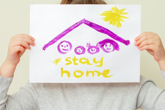 Zbliżenie dłoni dziecka trzyma zdjęcie sylwetki rodziny pod dachem i słowa „Zostań w domu” zakrywające jej twarz. Dzieci w koncepcji kwarantanny.