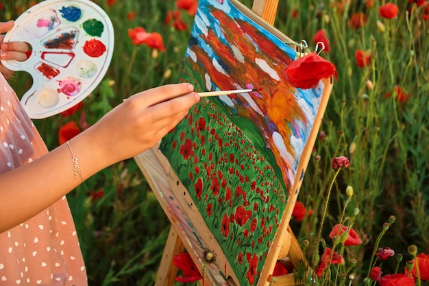 Zdjęcie zbliżenie dłoni dziecka z pędzlem i paletą farb w polu czerwonych maków i malowanie na płótnie