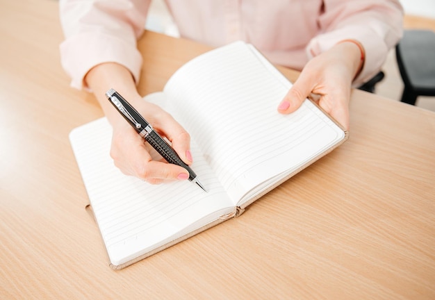 Zbliżenie dłoni kobiety piszącej w notatniku umieszczonym na drewnianym stole w biurze domowym oraz planach i prognozach koncepcji pracy