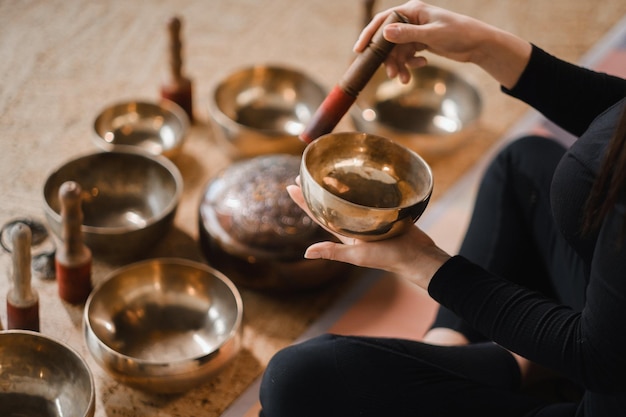 Zbliżenie dłoni kobiety siedzącej w pozycji lotosu przy użyciu misy śpiewającej w pomieszczeniu Relaks i medytacja Terapia dźwiękiem medycyna alternatywna Miski tybetańskie