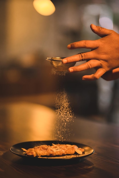 Zbliżenie dłoni młodego szefa kuchni posypującego cukrem pudrem na słodkie danie serwowane w czarnej płycie z orzechami