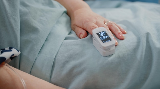 Zbliżenie dłoni wieku pacjenta z pulsoksymetrem w łóżku. Starsza osoba mająca narzędzie medyczne na palcu do pomiaru nasycenia tlenem i ciśnienia tętna. Emeryt z chorobą