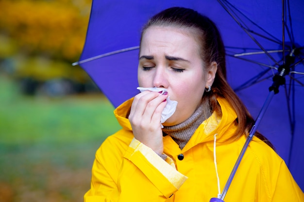 Zbliżenie dziewczyna jesienią z parasolką i płaszczem przeciwdeszczowym chora kobieta wydmuchuje nos i kicha na zewnątrz...
