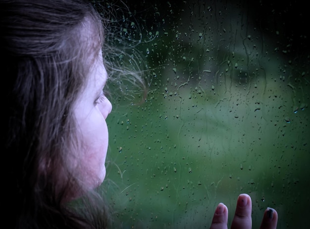Zdjęcie zbliżenie dziewczyny patrzącej przez mokre okno w porze deszczowej