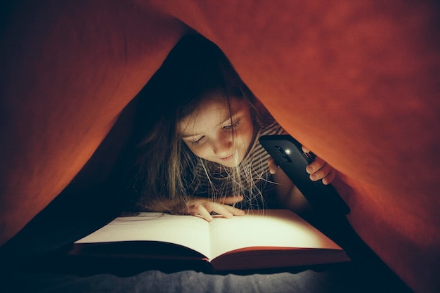 Zdjęcie zbliżenie dziewczyny z telefonem komórkowym czytającej książkę pod kocem