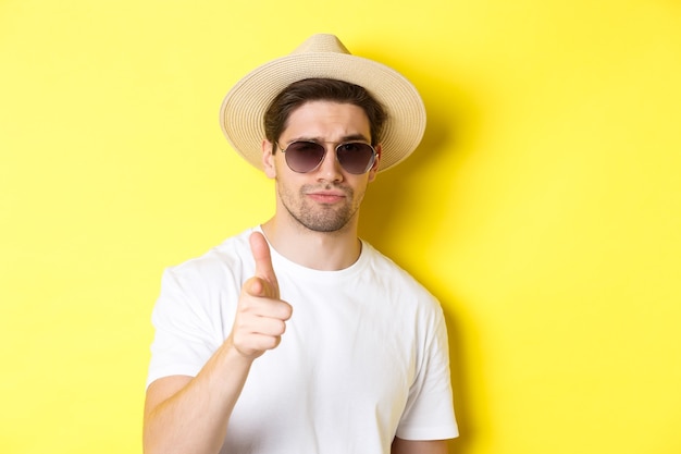 Zbliżenie: fajny facet w letnim kapeluszu i okularach przeciwsłonecznych, wskazując palcem z przodu, stojąc na żółtej ścianie