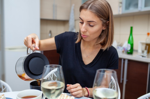 Zbliżenie fotografia kobiety nalewa herbaty w filiżankę. Na boku na stole jest kieliszek wina i kawałek ciasta