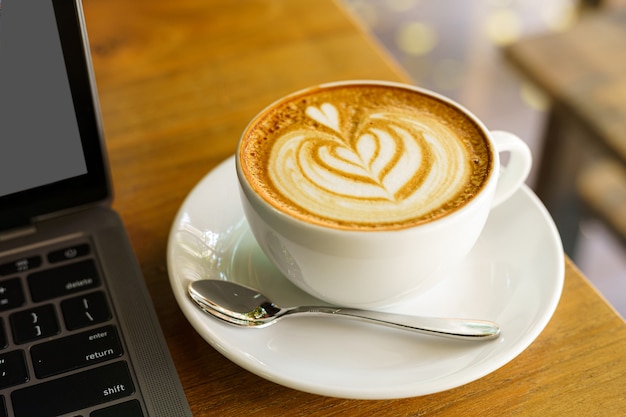 Zbliżenie: gorąca kawa latte z pianką mleczną latte art w filiżance kubek i laptop na drewniane biurko biurko w kawiarni w kawiarni, podczas koncepcji pracy biznesowej