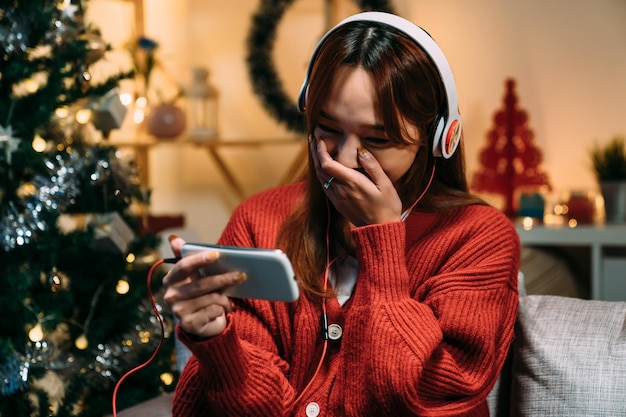 zbliżenie japońska kobieta trzymająca smartfon zakrywa usta, śmiejąc się ze śmiesznych filmów online ze słuchawkami w świątecznym salonie w wigilię Bożego Narodzenia
