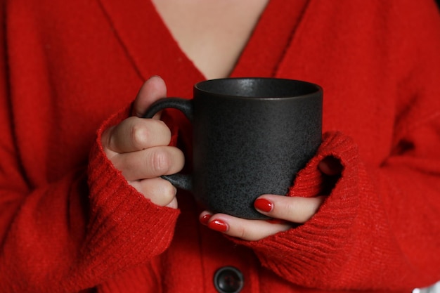 zbliżenie kobiecych rąk trzymających szarą filiżankę kawy, ubranych w czerwony ciepły, przytulny sweter