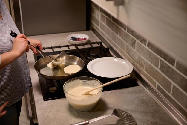 Zdjęcie zbliżenie: kobieta kucharz, gospodyni domowa nalewająca okrągłe ciasto płynne i gotująca naleśniki na patelni, uśmiecha się słodko, stojąc w pobliżu czarnego pieca w domowej kuchni. koncepcja ostatki wtorek