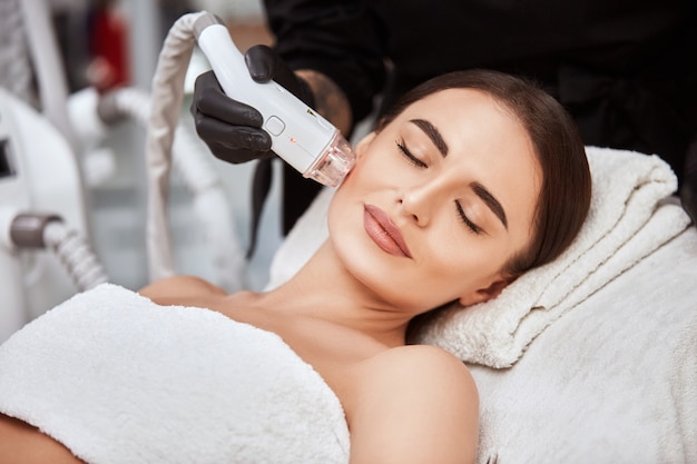 Zbliżenie kobiety otrzymującej zabieg na twarz laserem przez kosmetyczkę w salonie spa, przyjemna kobieta leżąca z zamkniętymi oczami i poddająca się terapii twarzy