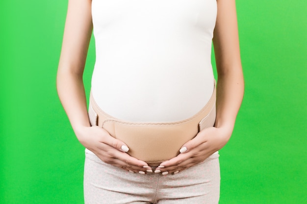 Zbliżenie kobiety w ciąży noszenia gorsetu ciążowego z bólem pleców na zielonym tle z miejsca na kopię. Koncepcja ortopedycznego pasa brzusznego.