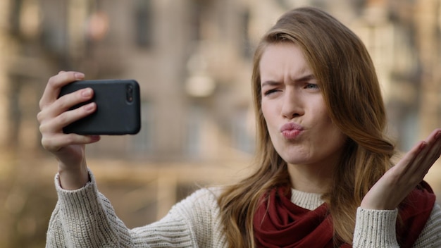 Zbliżenie krzywiąc się dziewczyna robi zdjęcie selfie smartfonem na ulicy miasta Portret wesołej hipsterki robiącej miny aparatem telefonu komórkowego na zewnątrz Zabawna dziewczyna patrząca na ekran telefonu komórkowego na ulicy