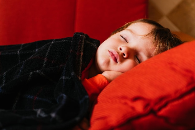 Zbliżenie małego kaukaskiego chłopca śpiącego na czerwonej kanapie przykrytej kocem