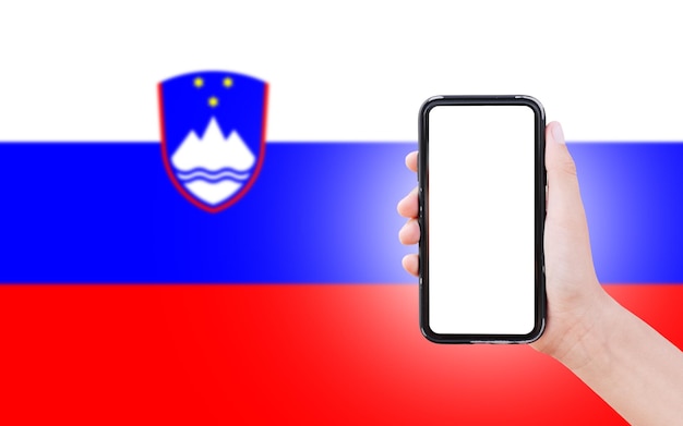 Zbliżenie męskiej dłoni trzymającej smartfon z pustym ekranem na tle niewyraźnej flagi Słowenii