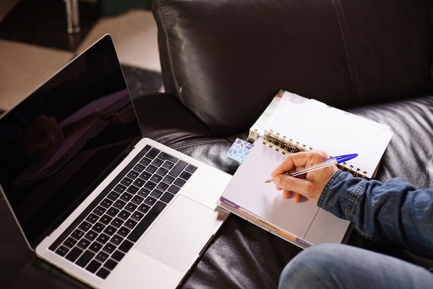 Zbliżenie męskiej dłoni z piórem zapisując notatki w zeszycie i używając laptopa na kanapie w domu