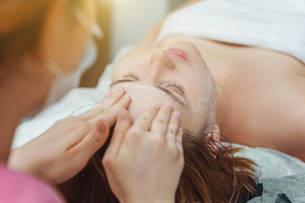 Zdjęcie zbliżenie: młoda piękna brunetka kobieta coraz zabieg na twarz z białym odżywczym kremem w salonie spa. masaż twarzy. spa do pielęgnacji skóry i ciała. pielęgnacja skóry oczyszczająca kosmetyczna koncepcja spa relaks