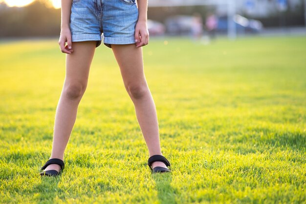 Zbliżenie młode dziecko dziewczyna nogi w dżinsowe szorty, stojąc na zielonej trawie trawnik na ciepły letni wieczór.