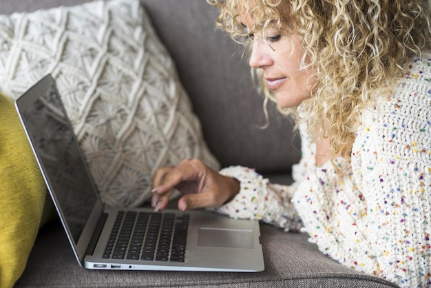 Zbliżenie młodej blondynki piszącej i używającej na laptopie w połączeniu bezprzewodowym online Nowoczesna kobieta pracująca na komputerze leżącym na kanapie w domu w samotności w czasie wolnym w pomieszczeniach Media społecznościowe
