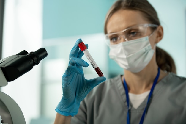 Zbliżenie: młody badacz laboratoryjny w masce i goglach stojący przed mikroskopem i trzymający próbkę krwi pacjenta z koronawirusem