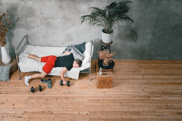 Zdjęcie zbliżenie: młody człowiek w mundurze sportowym odpoczywa, śpi na łóżku z hantlami. sportowiec odpoczywa i odkłada trening