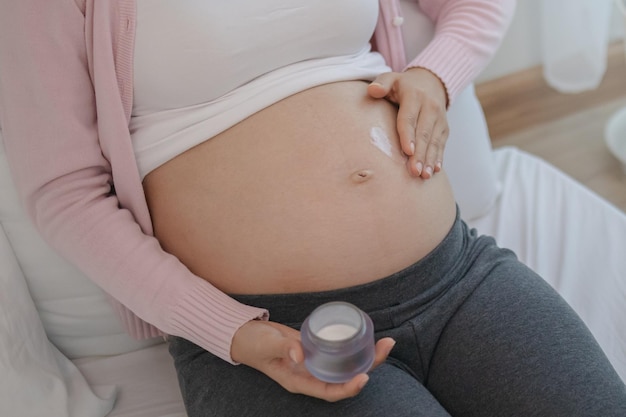 Zbliżenie na azjatyckie młode kobiety w ciąży stosujące balsam ciążowy, aby zapobiec rozstępom na brzuchu, wcierając krem nawilżający dla urody brzucha.