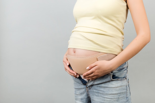 Zbliżenie na elastyczny zespół macierzyński ubrany na kobietę w ciąży, aby zmniejszyć ból pleców na szarym tle z miejsca na kopię. Koncepcja ortopedycznego pasa brzusznego.