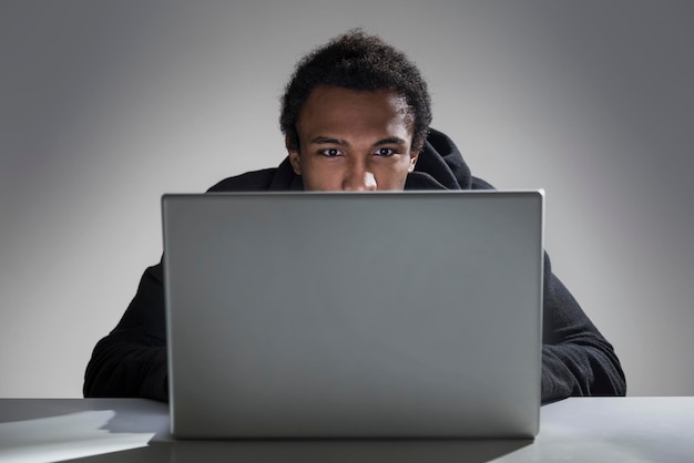 Zbliżenie na młodego Afroamerykanina w czarnej bluzie z kapturem, siedzącego przy stole i czytającego z ekranu laptopa. Makieta