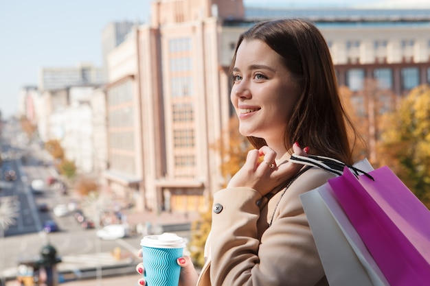 Zbliżenie na piękną szczęśliwą kobietę pijącą kawę, trzymającą torby na zakupy na zewnątrz