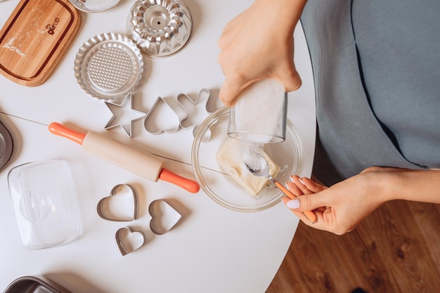 Zbliżenie na proces tworzenia plików cookie. Kobiece ręce mieszają składniki w misce. Do masła wsypać cukier. W pobliżu znajdują się foremki do ciastek i sprzęt kuchenny.