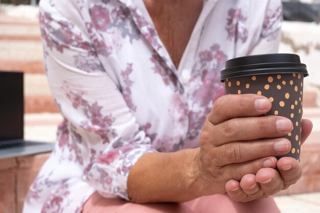 Zbliżenie na ręce starszej kobiety trzymając filiżankę kawy na wynos siedząc na zewnątrz na schodach w mieście w pobliżu jej laptopa