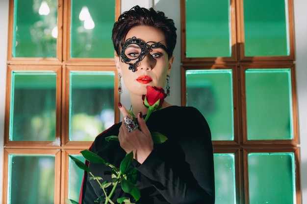 Zbliżenie na tajemniczą kobietę z czarną koronkową maską opartą na starych drzwiach i trzymającą czerwoną różę