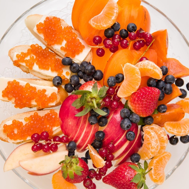 Zdjęcie zbliżenie na talerzu z asortymentem soczystych owoców organiczne jabłka mandarynki persimmon jagoda czerwona porzeczka truskawka i chleb z czerwonym kawiorem deser lub przekąska