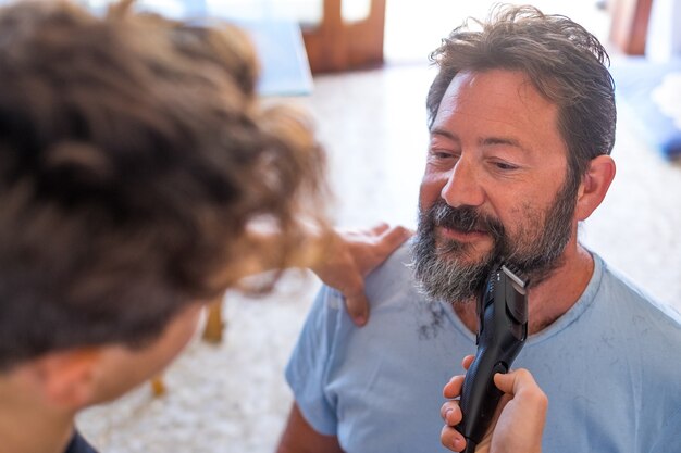 Zbliżenie nastolatka tnącego i czyszczącego brodę swojego taty w kwarantannie, aby być zabawnym i bawić się razem w domu - dwoje szczęśliwych ludzi w pomieszczeniu bawiących się w salonie fryzjerskim