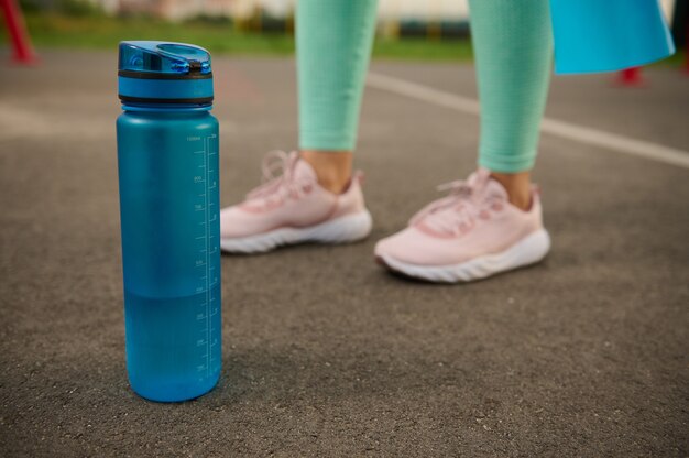 Zbliżenie niebieskiej plastikowej butelki ze słodką wodą na asfalcie boiska sportowego obok sportowych kobiecych stóp. Koncepcja pielęgnacji zdrowia i ciała