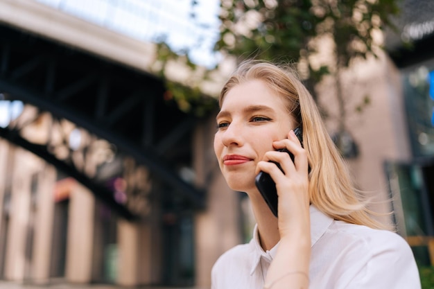 Zbliżenie niski kąt widzenia twarzy uroczej młodej kobiety rozmawiającej przez telefon komórkowy trzymającej smartfona blisko ucha, siedzącej samotnie na ławce na ulicy miasta