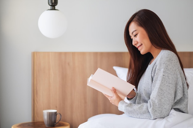 Zbliżenie obraz pięknej młodej azjatyckiej kobiety czytającej książkę w białym wygodnym łóżku w domu
