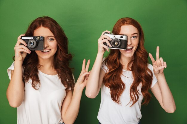 Zbliżenie obrazu dwóch rudowłosych kobiet 20s fotografujących cię aparatem retro i gestykulujących znak zwycięstwa, odizolowane na zielonym tle