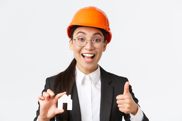 Zbliżenie optymistycznej profesjonalnej kobiety azjatyckiej inżyniera, architekta w kasku i garniturze zachęca do skontaktowania się z jej firmą w celu renowacji i projektowania, pokazując dom i kciuk w górę