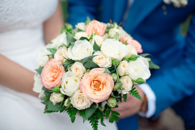 Zbliżenie pana młodego w garniturze i panna młoda w białej sukni stoją i trzymają bukiet brzoskwiniowych róż, eustom i kwiatów oraz zieleni