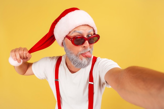Zbliżenie podekscytowany szary Brodaty mężczyzna w czerwonym kapeluszu Świętego Mikołaja wygłupiać się, pokazując język, co selfie na aparacie prowadzenie bloga Kryty studio strzał na białym tle na żółtym tle
