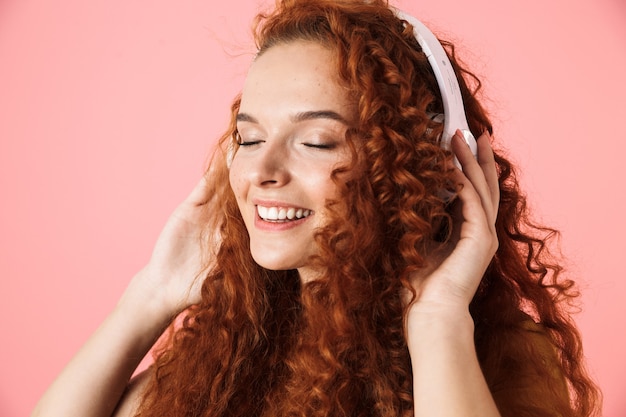 Zbliżenie Portret Atrakcyjnej Uśmiechniętej Młodej Kobiety Z Długimi Kręconymi Rudymi Włosami Stojącej Na Białym Tle, Słuchającej Muzyki Przez Słuchawki