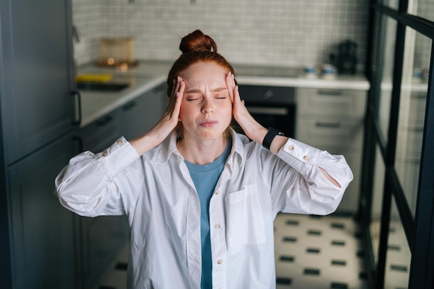 Zbliżenie portret cierpiącej rudej młodej kobiety masującej skronie zmniejszające ból głowy w domu Przytłoczona zmęczona tysiącletnia biznesowa dama cierpiąca na wysokie ciśnienie krwi lub migrenę