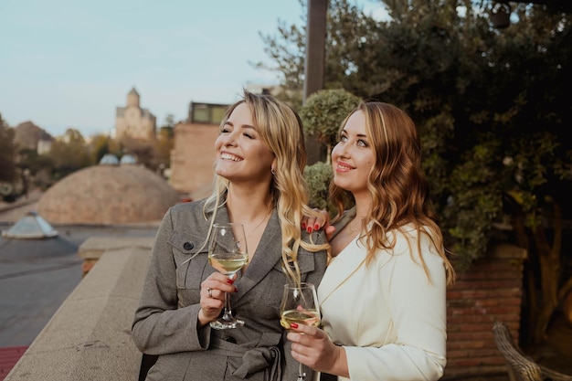 Zbliżenie portret dwóch koleżanek w surowych garniturach, śmiejąc się, pijąc wino na tarasie na zewnątrz