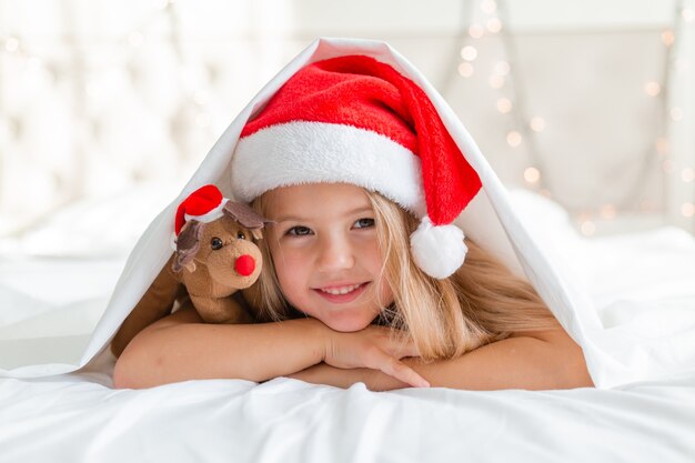 zbliżenie portret małej blondynki leżącej w łóżku w kapeluszu Świętego Mikołaja z jelonkiem-zabawką