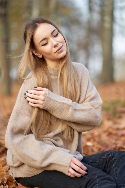 Zbliżenie portret piękna stylowa blondynka pewna siebie dziewczyna z zamkniętymi oczami i długie blond włosy sobie przytulny sweter siedzi w parku jesień.